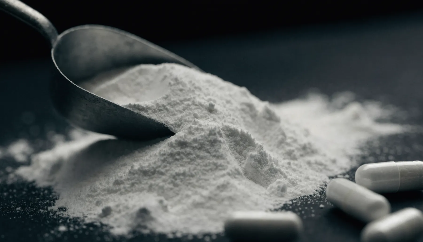 Supplement powder spilling from a metal scoop beside loose unbranded capsules